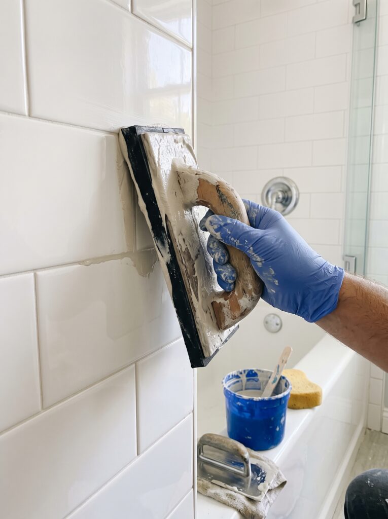 Gloved hand pressing grout into a tile joint with a rubber float held at an angle beside a bucket of grout