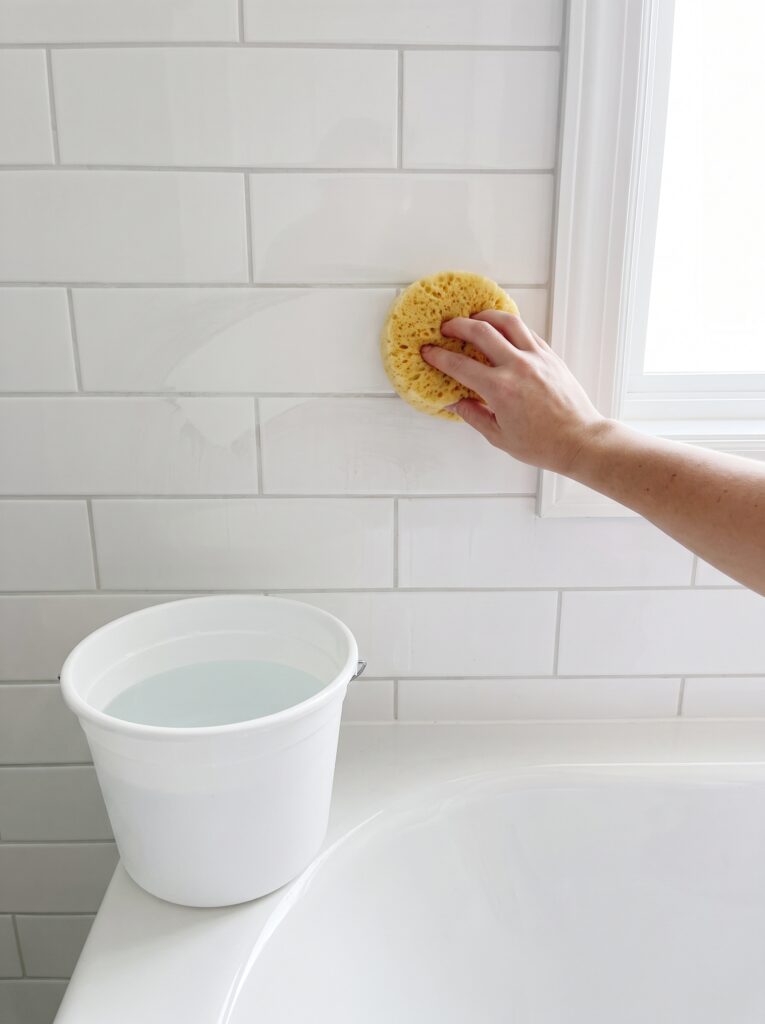 Hand wiping a damp yellow sponge across a white subway tile wall beside a white bucket of clear water on a tub ledge