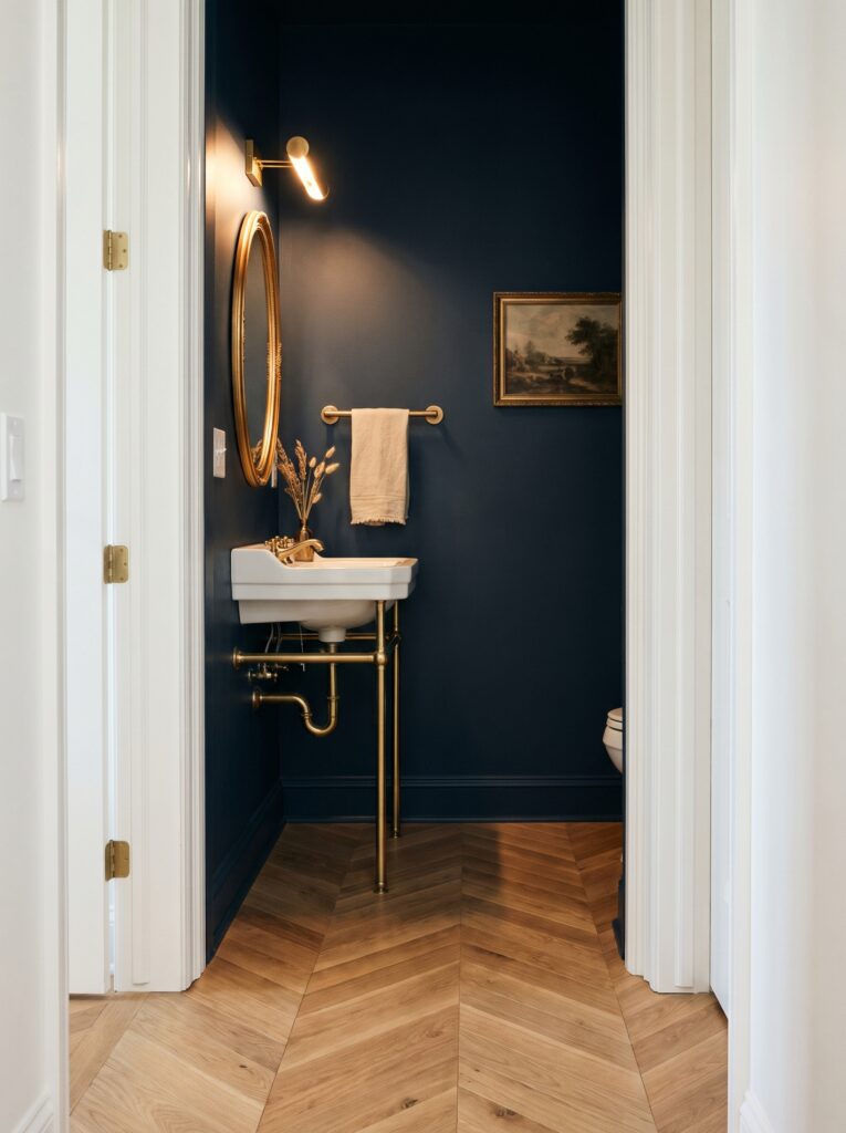 engineered oak chevron floor running from the hallway into a navy half bath with a brass console sink