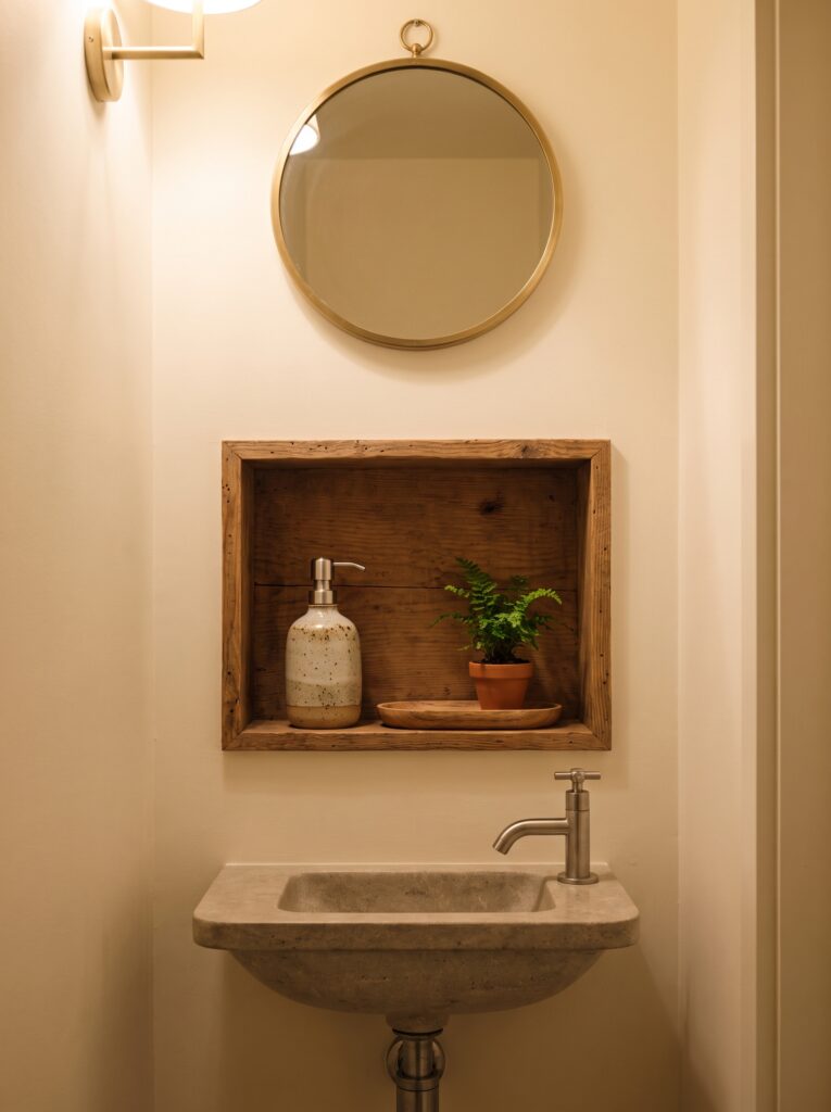 A small wood-framed niche above a stone wall-hung sink holding a soap pump, a fern and a wooden tray, with a round mirror above