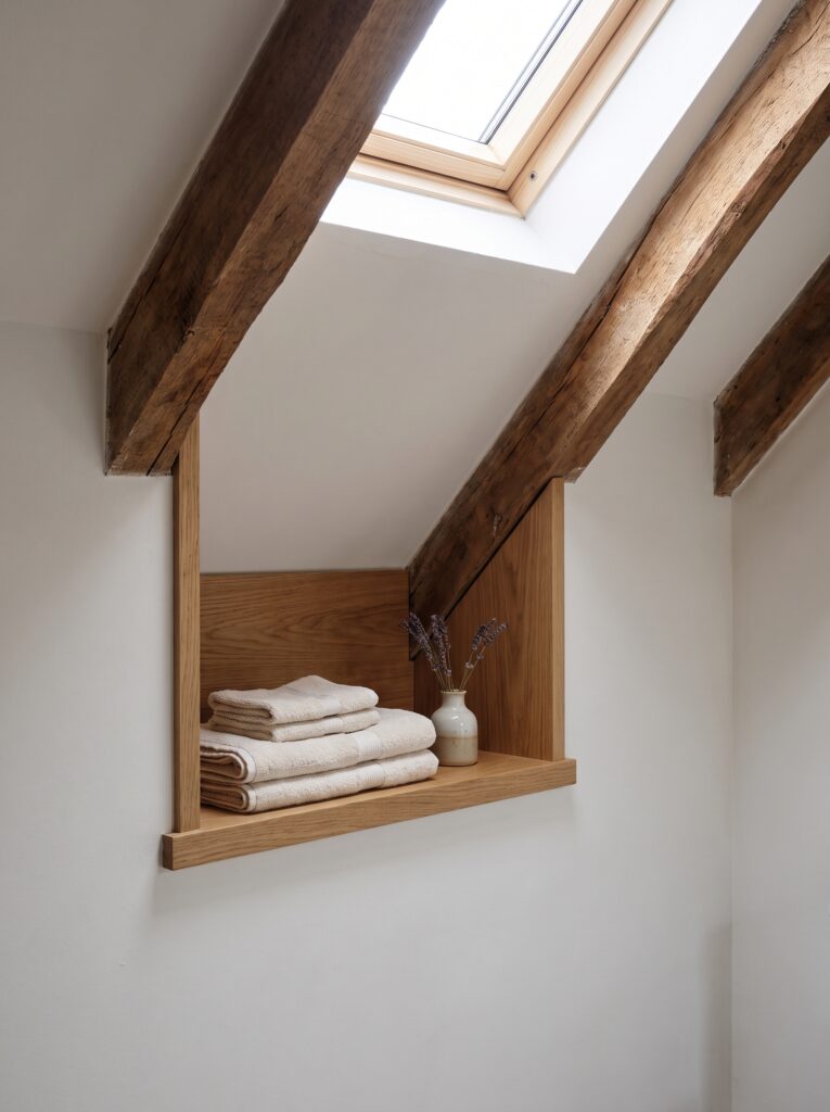 An oak-lined niche built between rafters under a sloped attic ceiling beside a skylight, holding folded towels and a small vase of lavender