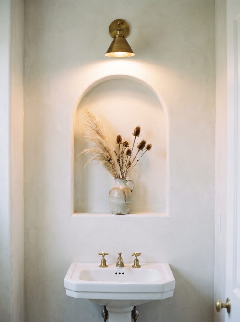 An arched plaster niche above a pedestal sink in a limewashed powder room, lit by a brass sconce and holding a vase of dried stems
