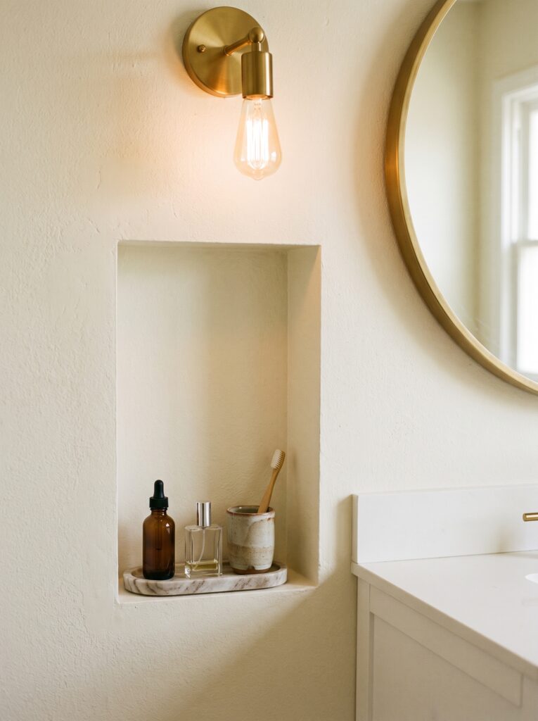 A tall recessed niche in a cream plaster wall beside a round brass mirror, holding a marble tray with an amber dropper bottle, a perfume bottle and a toothbrush cup