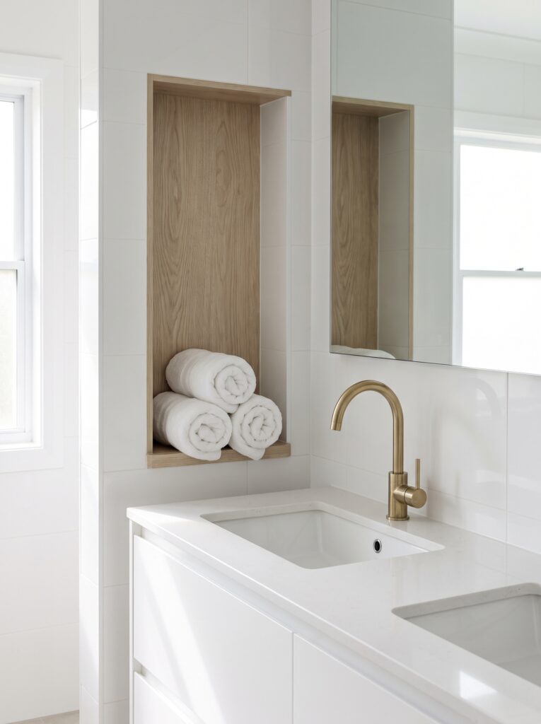 An oak-lined niche beside a white vanity holding three rolled white towels, next to a brass faucet and a mirror