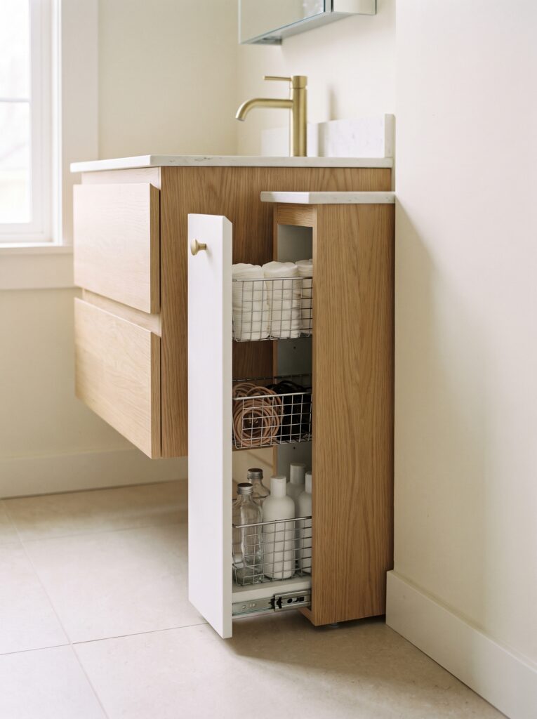 A slim white pull-out cabinet with three wire baskets holding cotton rounds, hair ties and plain bottles in the gap beside a floating oak vanity with a brass tap