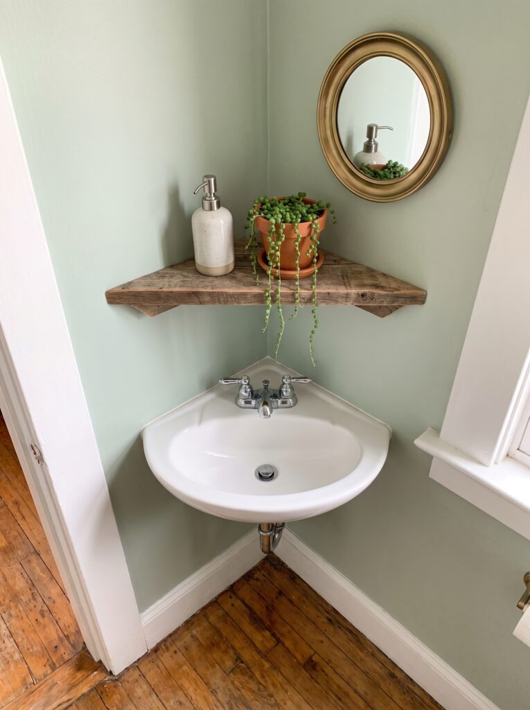 A wall-mounted corner sink in a sage green powder room with a triangular wooden shelf holding a soap pump and a plant, and a round brass mirror above