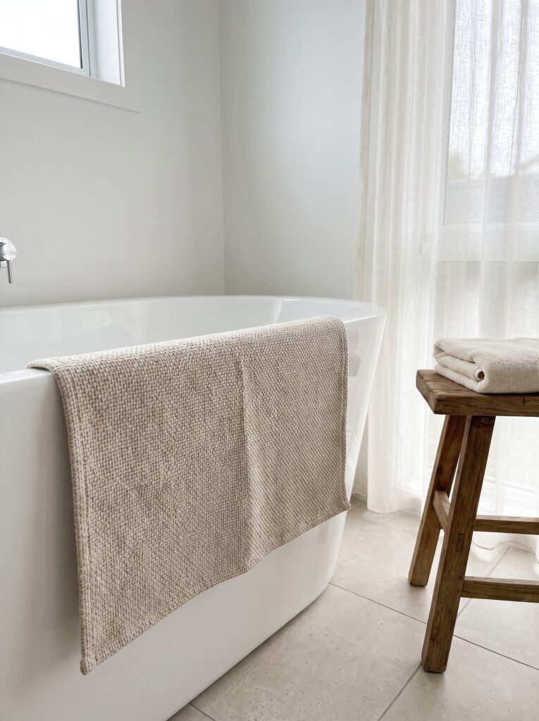 A beige woven bath mat draped over the side of a white freestanding tub to dry