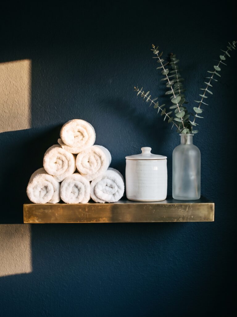 brass-edged shelf on a navy wall with a pyramid of rolled white towels, a white jar and a frosted vase of eucalyptus