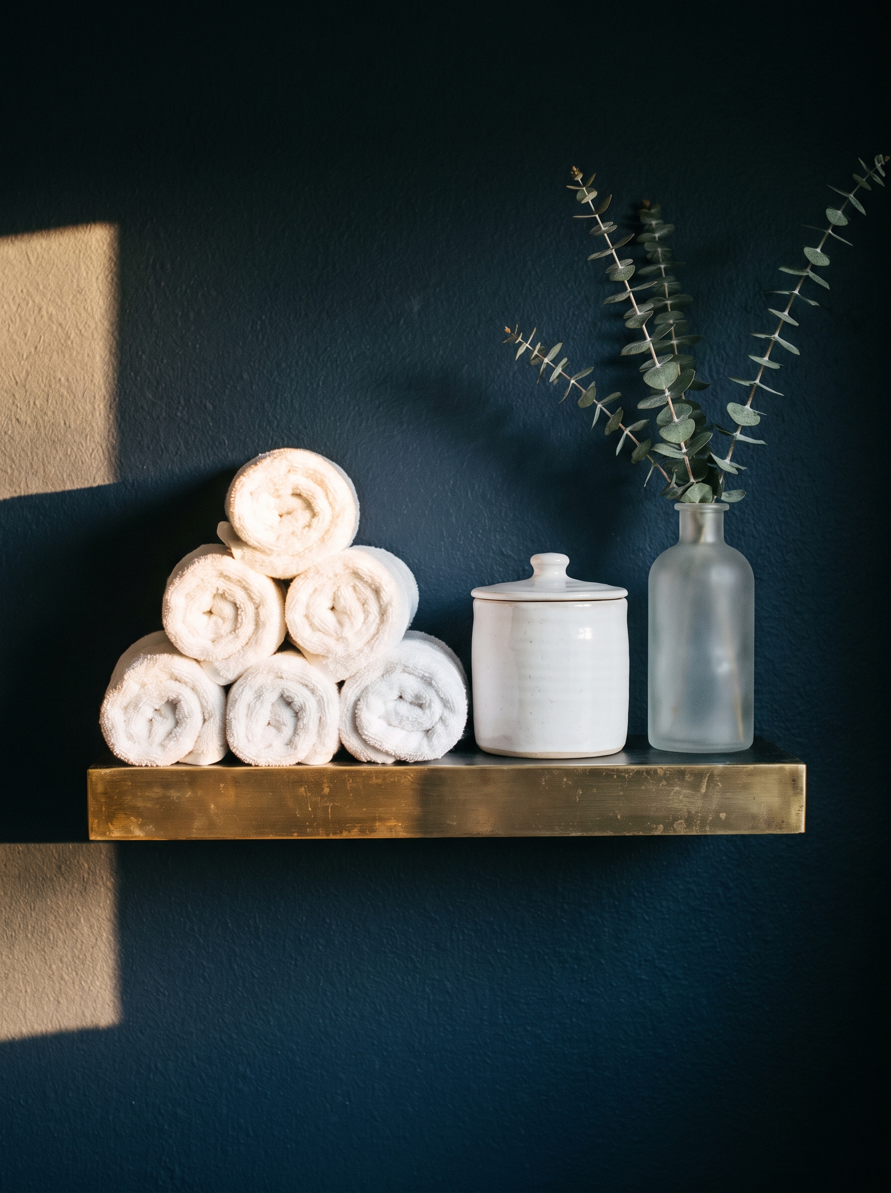 brass-edged shelf on a navy wall with a pyramid of rolled white towels, a white jar and a frosted vase of eucalyptus
