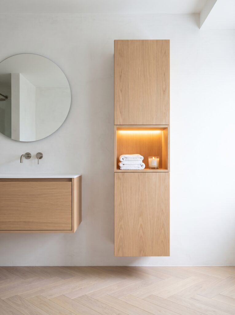 A wall-hung oak tower with a lit open niche holding a folded white towel and a candle beside a floating oak vanity and a round mirror.