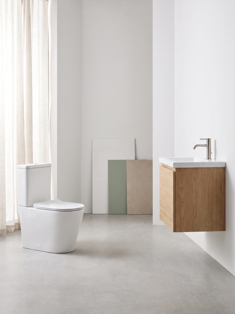 A bathroom showroom corner with a white toilet standing apart from a floating oak vanity with a brushed nickel tap, and three tile samples leaning on the wall