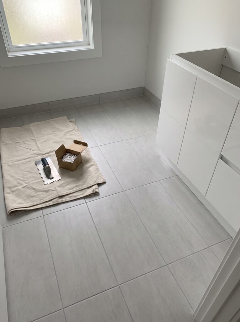 A newly tiled grey bathroom floor with a notched trowel and a small box on a folded drop cloth beside a new white vanity base under a frosted window