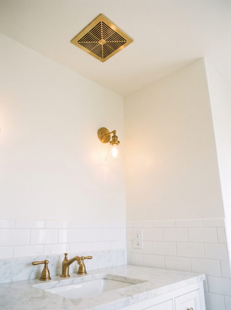 A brass ceiling exhaust fan grille above a white tiled bathroom wall with a brass sconce and a GFCI outlet beside a marble vanity with a brass faucet