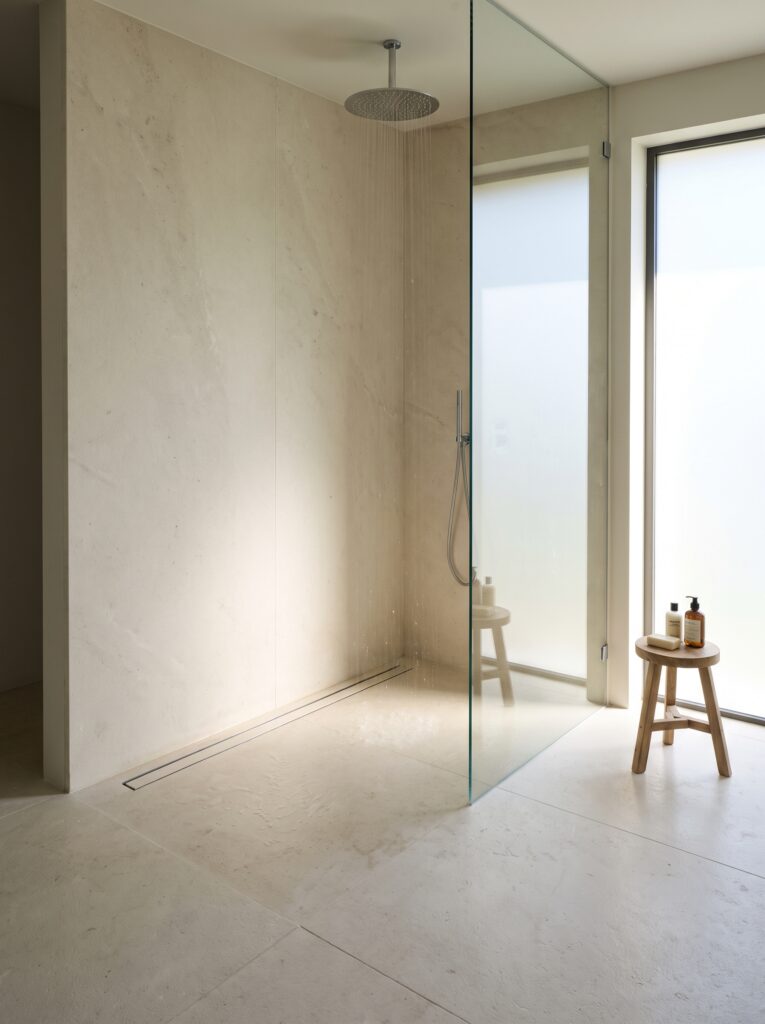 A curbless walk-in shower with a linear drain along the back wall, a stone floor and one fixed clear glass panel
