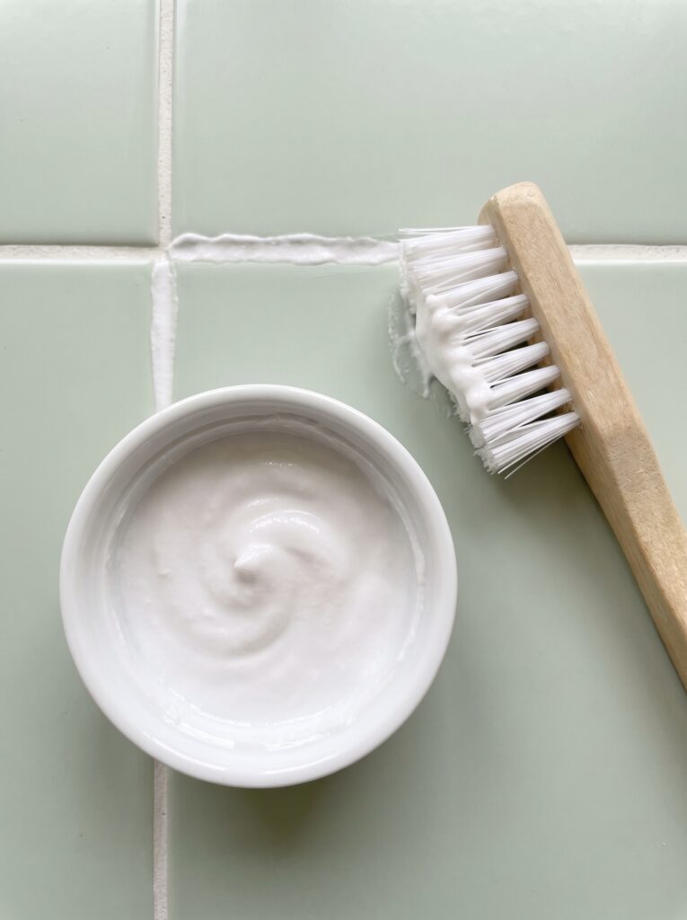 A white bowl of baking soda paste beside a nylon scrub brush with a line of paste in the grout joint between pale green tiles