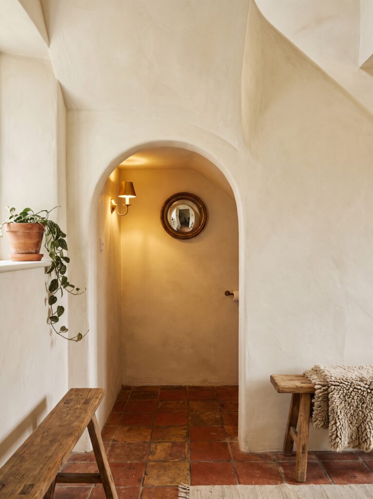 Plastered arched opening under a staircase leading to a terracotta-floored toilet with a brass sconce and a convex mirror.