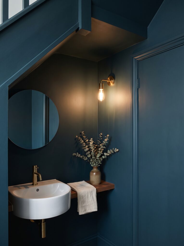 Deep teal walls, slope and door painted one color around a wall-hung basin, a round mirror and a brass bulb sconce.