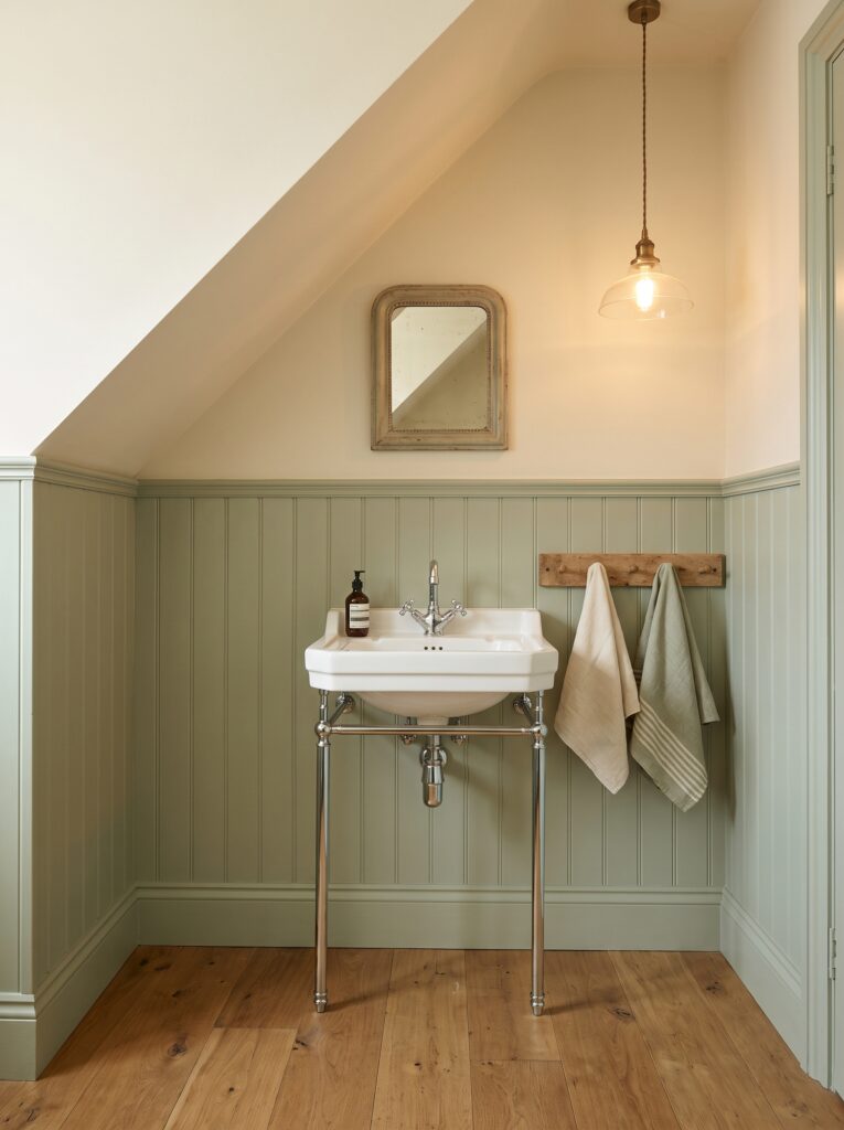 Half-height sage tongue and groove panelling under a sloped ceiling with a console basin on chrome legs and a peg rail with two towels.