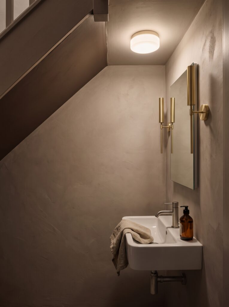 Under stairs powder room lit by a flush ceiling light and two brass sconces beside a tall rectangular mirror above a wall-hung basin.