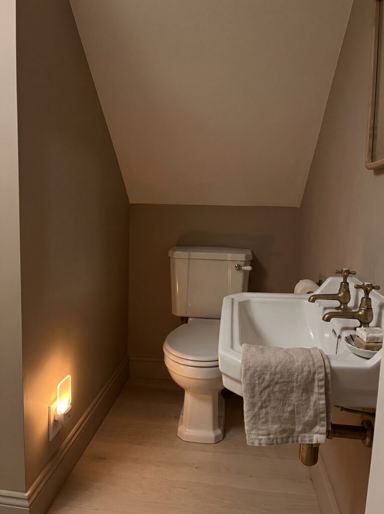 Amber plug-in night light glowing on the baseboard of a taupe under stairs toilet with a wall-hung basin and brass taps.