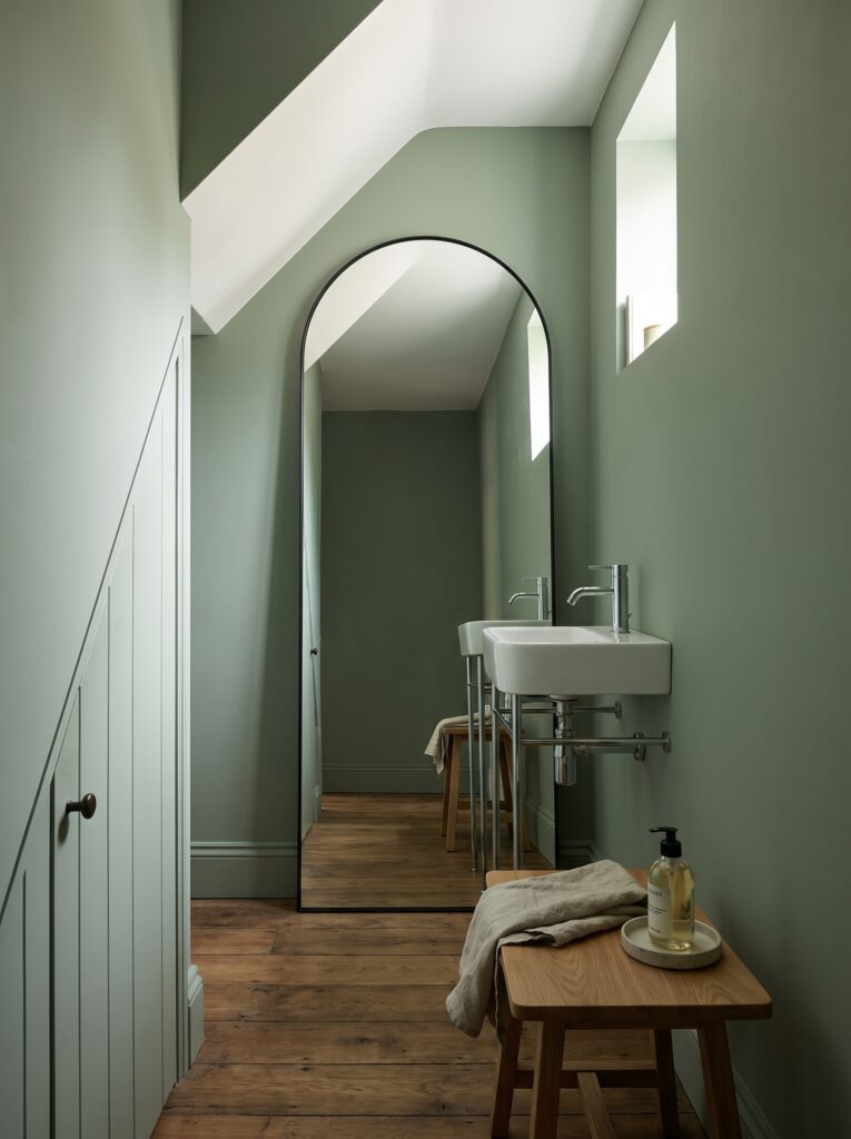 Tall black-framed arched mirror leaning in a sage green room beside a console basin and an oak stool with a towel.