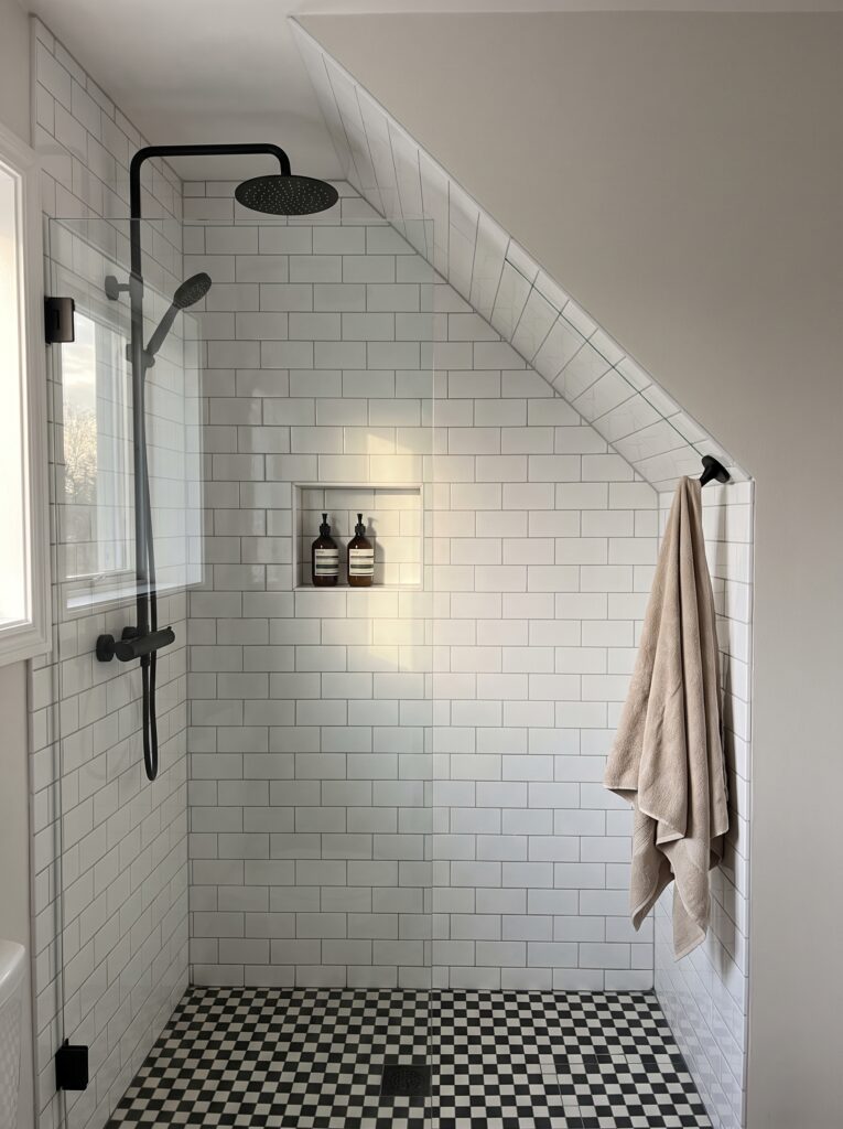 Walk-in shower under a sloped ceiling with white metro tiles, a glass panel, a black rain shower and a checkerboard floor.