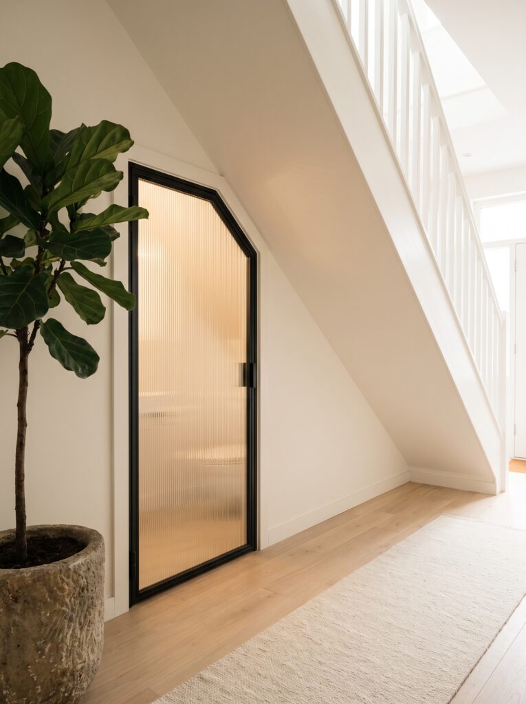Reeded glass door in a black steel frame with an angled top under a white staircase, lit warmly from inside, beside a fiddle leaf fig.