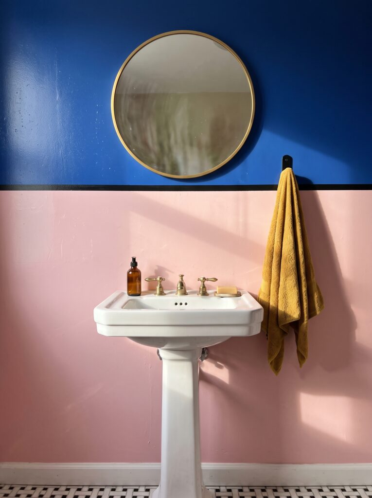 Bathroom wall split into glossy cobalt blue above and soft pink below with a thin black line, a round gold mirror, a white pedestal sink and a mustard towel on a black hook.