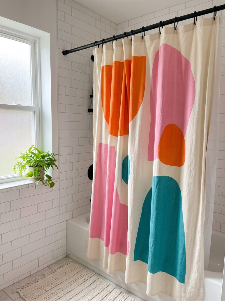 Large abstract shower curtain in orange, pink and teal shapes on natural cotton, hung on a black rod against white subway tile with a frosted window.