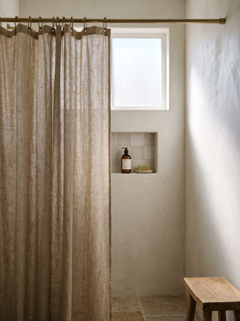 linen shower curtain on metal rings beside tiled shower niche with soap and shampoo