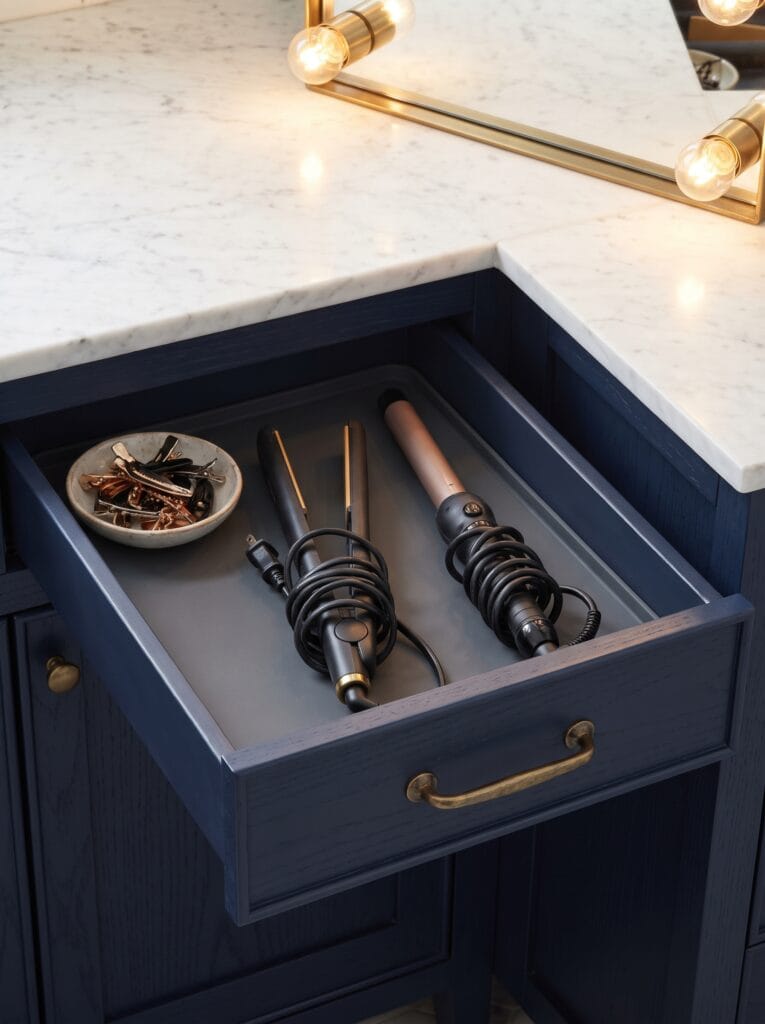 Navy bathroom vanity drawer lined with a grey heat resistant mat holding a flat iron and curling wand with coiled cords