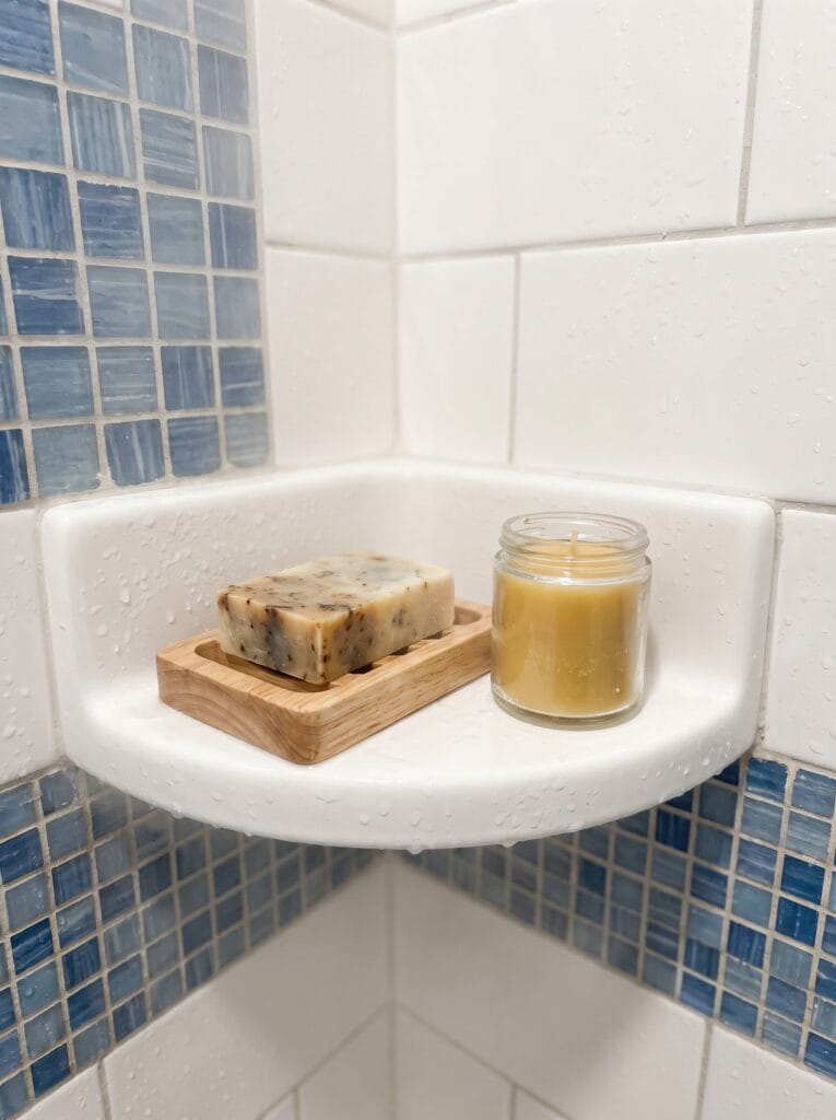 white corner shower shelf holding a bar of soap on a wood dish and a glass jar candle