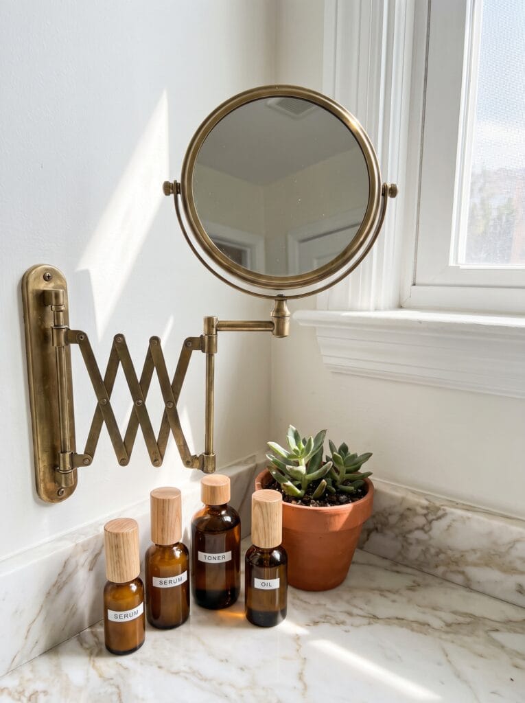 brass accordion extending mirror above four labeled amber glass skincare bottles on a marble counter