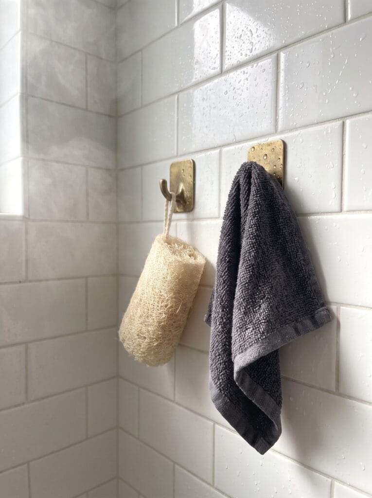 brass hooks in a wet shower holding a natural loofah and a charcoal gray washcloth