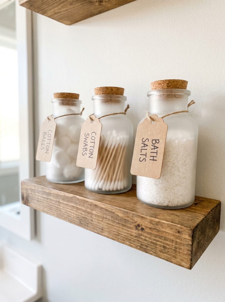 three frosted glass apothecary jars with cork lids holding cotton balls, cotton swabs, and bath salts