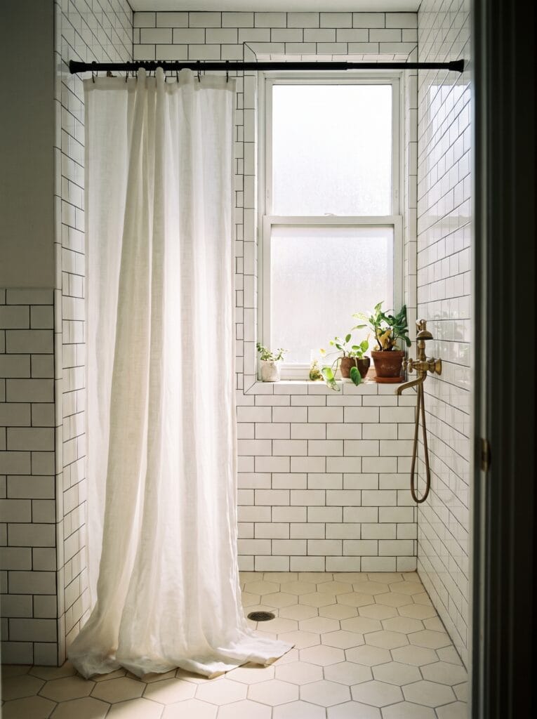 white linen shower curtain on a black tension rod in a tiled walk-in shower with plants on the windowsill