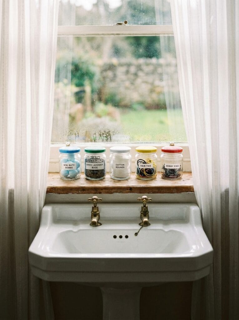 Five small jars with different colored lids lined up on a wood windowsill above a pedestal sink, sorted by bath bombs, dried lavender, cotton rounds, hair ties, and bobby pins