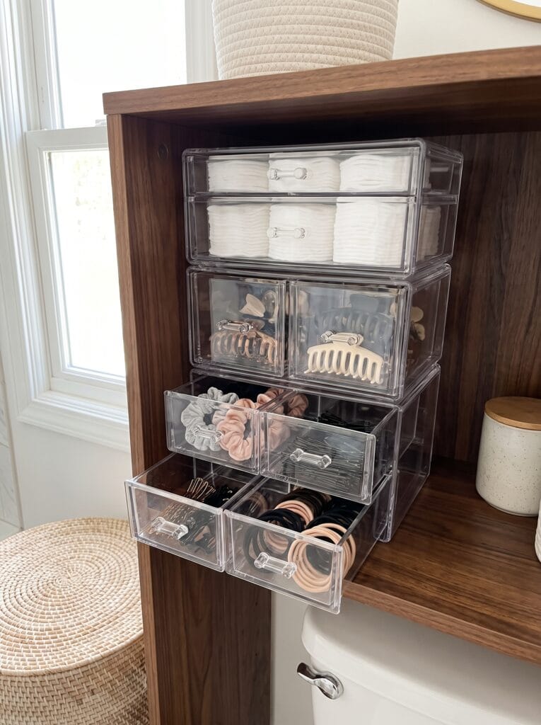 clear stackable drawer organizers holding cotton pads, hair clips, scrunchies, and hair ties on a bathroom shelf
